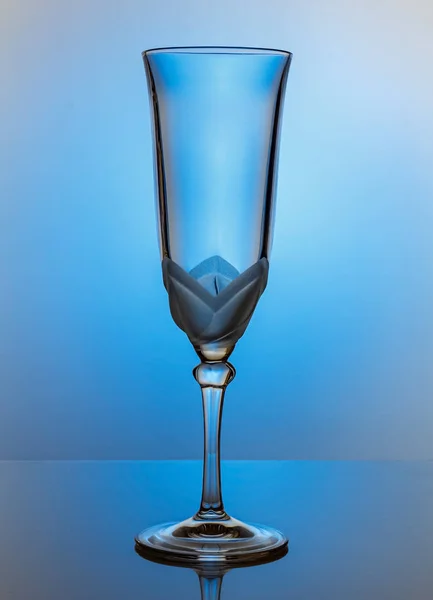 Empty transparent champagne glass on a thin high leg in light blue gradient backlight on a glossy surface — Stock Photo, Image