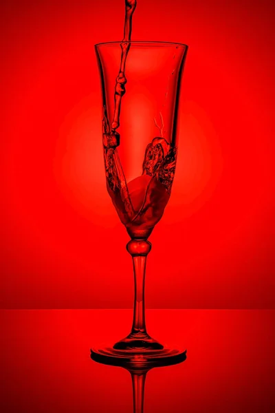 Замороженные струи воды с брызгами в пустой прозрачный бокал шампанского на тонкой высокой ноге в ярко-красном фоне с отражением на темно-глянцевой поверхности. Стоп-моция — стоковое фото