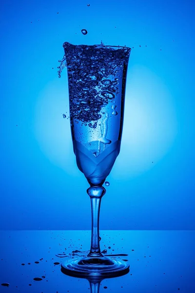 Chorro de agua congelada con salpicaduras y gotas de agua volando en copa de champán completa en una pierna alta delgada con reflejo en una superficie brillante en la luz de fondo azul. Detener fotografía en movimiento — Foto de Stock