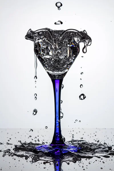 Pełna szklanka koktajlowa na niebieskiej cienkiej i wysokiej nodze z zamarzniętymi strumieniami wody i kroplami wody na ciemnej, błyszczącej powierzchni w białym podświetleniu. Stop fotografii ruchu — Zdjęcie stockowe