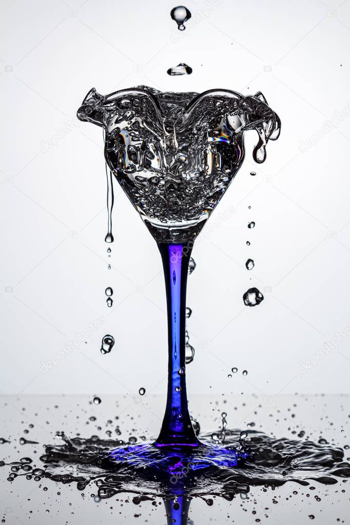 Full cocktail glass on blue thin and high leg with frozen water streams and flying water drops on dark glossy surface in white backlight. Stop motion photography