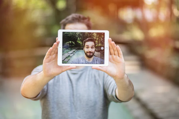 Joven guapo haciendo una cara linda tonta mientras toma una selfie con una tableta. graduado con una llamarada —  Fotos de Stock