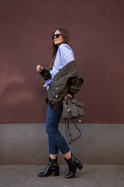 Traje de moda de primavera perfecto. blogger europeo de moda con una blusa de rayas crossover de moda con un nudo frontal, una chaqueta de bombardero, jeans, botas de tobillo y sosteniendo un bolso con estilo . —  Fotos de Stock