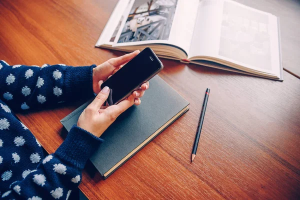 Young girl in a sweater holding a smart phone in hands on the background of books on the table