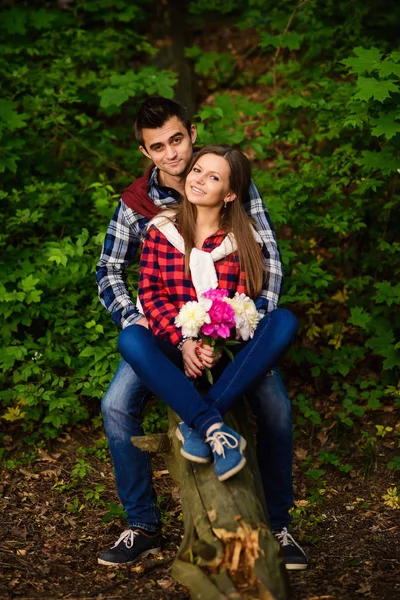 Stylish young couple in shirts and jeans while walking in the forest. A beautiful girl with her handsome boyfriend sitting on a fallen tree.