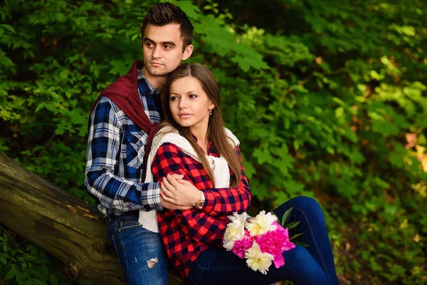 Snygga unga par i skjortor och jeans medan promenader i skogen. En vacker flicka med sin stiliga pojkvän i en kram som sitter på ett fallna träd. — Stockfoto