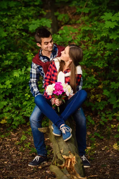 Stylish young couple in shirts and jeans while walking in the forest. A beautiful girl with her handsome boyfriend sitting on a fallen tree.