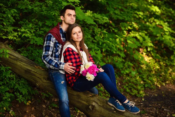 Snygga unga par i skjortor och jeans medan promenader i skogen. En vacker flicka med sin stiliga pojkvän som sitter på ett fallna träd. — Stockfoto