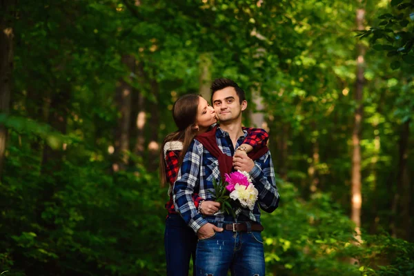 Elegante jovem casal em camisas e jeans enquanto caminhava na floresta. Uma menina encantadora beija na bochecha seu namorado bonito . — Fotografia de Stock