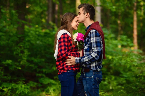 Elegante jovem casal em camisas e jeans enquanto caminhava na floresta. Um namorado bonito beija sua bela namorada encantadora na testa . — Fotografia de Stock