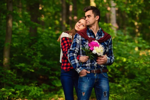 Stylish young couple in shirts and jeans while walking in the forest. The charming girl with bouquet of flowers laid her head on the shoulder of her handsome boyfriend.