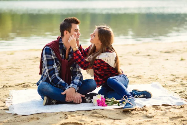 Jeune couple élégant en chemises et jeans tout en marchant le long de la rive du lac. Une belle fille attrayante et son beau petit ami assis sur une couverture et souriant . — Photo