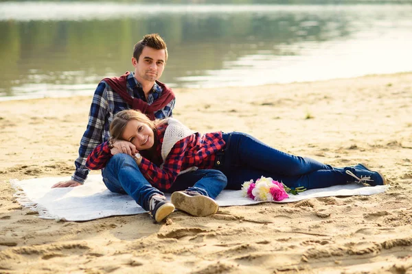 Elegante giovane coppia in camicie e jeans mentre cammina sulla riva di un lago. Una bella ragazza attraente sdraiata in ginocchio al suo ragazzo, seduta su un plaid . — Foto Stock