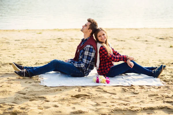 Stylish young couple in shirts and jeans while walking on the shore of a lake. Beautiful attractive girl and her handsome boyfriend leans to each other sitting on a plaid.