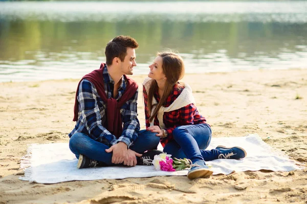 Elegante pareja joven en camisas y jeans mientras camina a lo largo de la orilla del lago. Una hermosa chica atractiva y su novio guapo sentados en una manta y sonriendo el uno al otro . — Foto de Stock