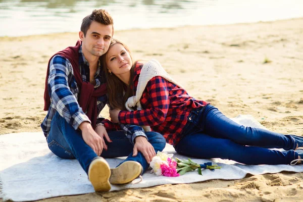 Snygga unga par i skjortor och jeans medan promenader på stranden av en sjö. Vackra attraktiva flicka och hennes stiliga pojkvän lutar till varandra sitter på en pläd. — Stockfoto