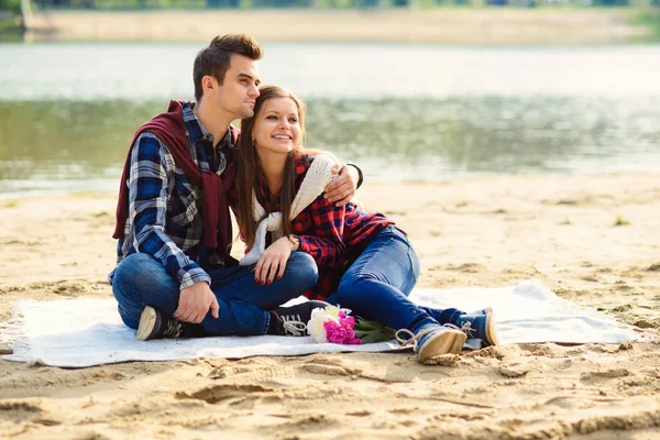 Elegante joven pareja sonriente en camisas y jeans mientras camina en la orilla de un lago. Hermosa chica atractiva y su novio guapo se inclina el uno al otro sentado en un cuadros . — Foto de Stock