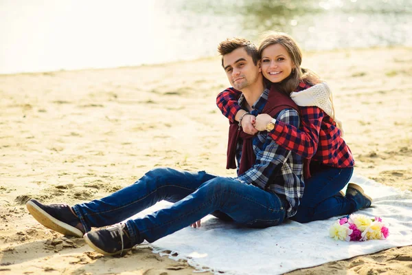 Snygga unga par i skjortor och jeans medan promenader på stranden av en sjö. Vackra attraktiva flicka kramar sin stiliga pojkvän som sitter på en pläd. — Stockfoto
