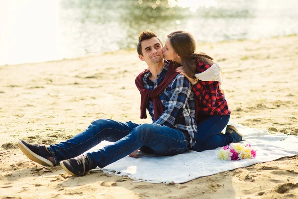 Elegante pareja joven en camisas y jeans mientras camina en la orilla de un lago. Hermosa chica atractiva abraza y besa en la mejilla a su novio guapo sentado en un cuadros . — Foto de Stock