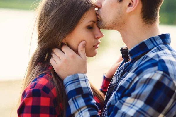 Elegante pareja joven en camisas y jeans mientras camina a lo largo de la orilla del lago. Un novio guapo besa a su hermosa novia encantadora en la frente. De cerca. . — Foto de Stock