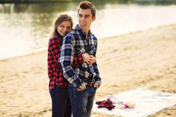 Snygga unga par i skjortor och jeans medan promenader på stranden av en sjö. Vackra charmiga flicka kramar sin stiliga pojkvän. — Stockfoto