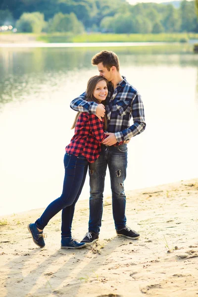 Elegante joven pareja sonriente en camisas y jeans mientras camina a lo largo de la orilla del lago. Una hermosa chica encantadora y su apuesto novio se apoyaron uno contra el otro . — Foto de Stock