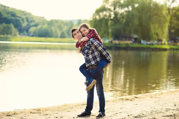 Elegante joven pareja sonriente en camisas y jeans mientras camina a lo largo de la orilla del lago. Hermosa chica encantadora saltó de nuevo a su novio guapo . — Foto de Stock
