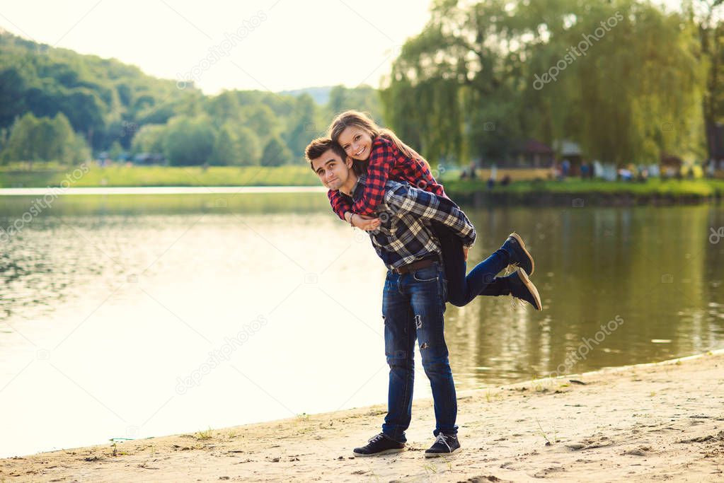 Stylish young couple in shirts and jeans while walking along the lake shore. Beautiful charming girl jumped on back to her handsome boyfriend.
