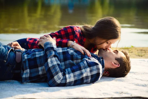 Elegante pareja joven en camisas y jeans mientras camina a lo largo de la orilla del lago. Una hermosa chica atractiva y su guapo novio acostado en una manta y sonriendo el uno al otro . — Foto de Stock