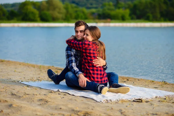Snygga unga par i skjortor och jeans medan promenader på stranden av en sjö. Vackra charmiga flicka kramar sin stiliga pojkvän som sitter på en pläd. — Stockfoto