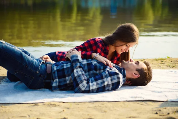 Elegante giovane coppia in camicie e jeans mentre passeggia lungo la riva del lago. Una bella ragazza attraente e il suo bel ragazzo sdraiato su una coperta e sorridente l'un l'altro . — Foto Stock