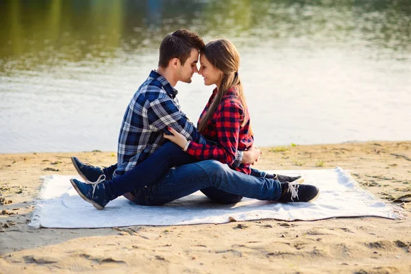 Elegante joven pareja sonriente en camisas y jeans mientras camina en la orilla de un lago. Hermosa chica atractiva y su novio guapo se inclina el uno al otro sentado en un cuadros . — Foto de Stock