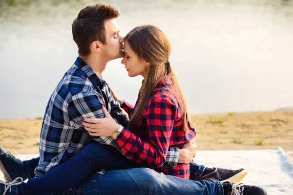 Elegante jovem casal sorridente em camisas e jeans enquanto caminhava na margem de um lago. Um namorado bonito beija sua bela namorada encantadora na testa sentada em uma xadrez . — Fotografia de Stock