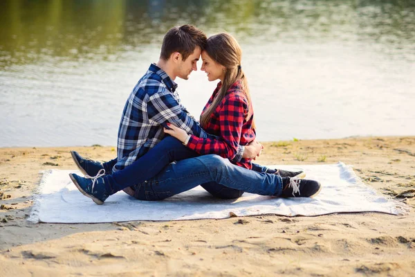 Stylish young smiling couple in shirts and jeans while walking on the shore of a lake. Beautiful attractive girl and her handsome boyfriend leans to each other sitting on a plaid. — Stock Photo, Image
