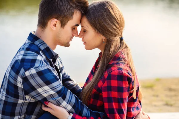 Stylish young smiling couple in shirts and jeans while walking on the shore of a lake. Beautiful attractive girl and her handsome boyfriend leans to each other sitting on a plaid. Close up. — Stock Photo, Image
