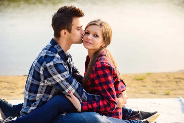 Elegante jovem casal sorridente em camisas e jeans enquanto caminhava na margem de um lago. Bonito namorado beijando na bochecha sua bela namorada encantadora sentado em uma xadrez . — Fotografia de Stock