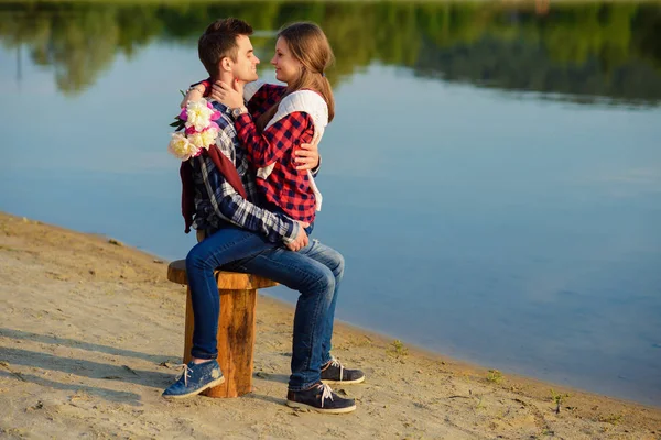 Stylish young couple in shirts and jeans sits on the stump on the shore of the lake. Beautiful attractive girl sitting on knees of her handsome boyfriend. — Stock Photo, Image