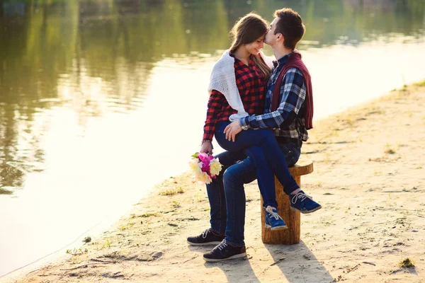 Casal jovem elegante em camisas e jeans senta-se no toco na margem do lago. Menina bonita atraente sentado de joelhos de seu namorado bonito . — Fotografia de Stock