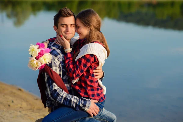 Elegante giovane coppia in camicie e jeans si siede sul ceppo sulla riva del lago. Bella ragazza attraente seduta sulle ginocchia del suo bel fidanzato . — Foto Stock