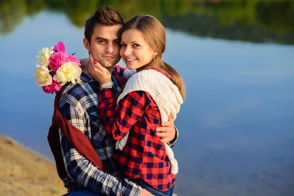 Stylish young couple in shirts and jeans sits on the stump on the shore of the lake. Beautiful attractive girl sitting on knees of her handsome boyfriend.