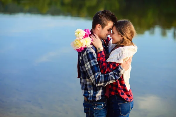 Stylish young smiling couple in shirts and jeans while walking on the shore of a lake. Beautiful attractive girl and her handsome boyfriend leans to each other on the background of the lake. — Stock Photo, Image