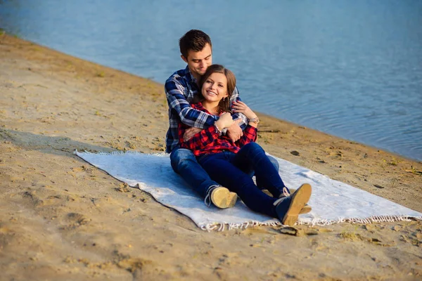 Elegante giovane coppia sorridente in camicie e jeans mentre cammina sulla riva di un lago. Bello fidanzato abbraccia la sua bella ragazza affascinante seduto su un plaid . — Foto Stock