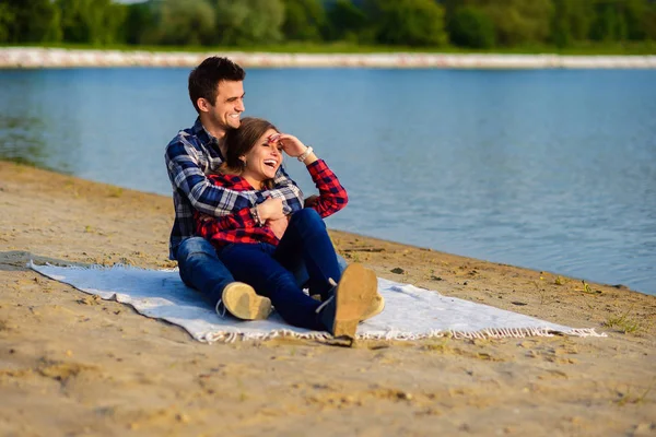 Snygg ung leende par i skjortor och jeans medan promenader på stranden av en sjö. Stiliga pojkvän kramar sin vackra charmiga flicka sitter på en pläd. — Stockfoto