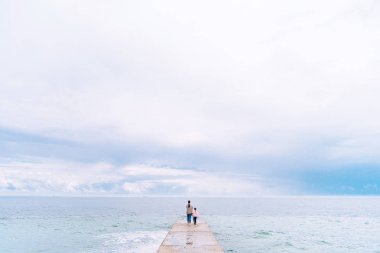 Anne küçük kızı olan kot pantolon ve t-shirt giymiş deniz ve gökyüzü arka plan üzerinde somut bir iskelede duran