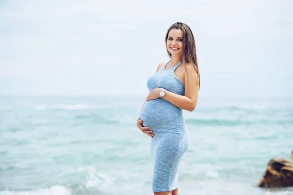 Μια όμορφη έγκυος γυναίκα σε ένα μπλε σφιχτό φόρεμα στην παραλία αγγίζοντας την κοιλιά της με αγάπη και φροντίδα. Περπάτημα στην παραλία, στην ηλιόλουστη μέρα. — Φωτογραφία Αρχείου