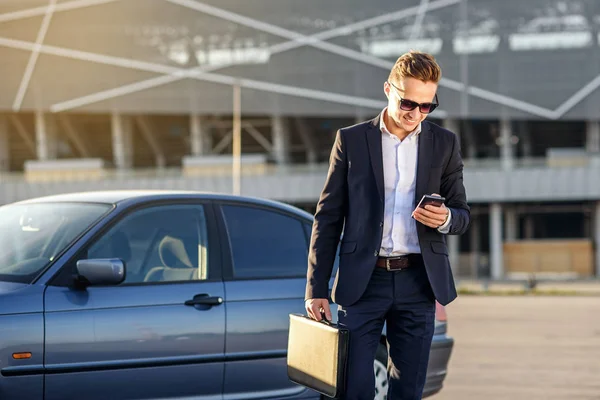 Empresário bonito atraente com diplomata e smartphone nas mãos perto do carro — Fotografia de Stock