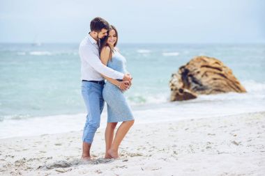 Beyaz bir gömlek ve Mavi pantolon cesur ve yakışıklı adam kayalar ve deniz arka planı mavi elbiseli güzel onun hamile eşi hugs.