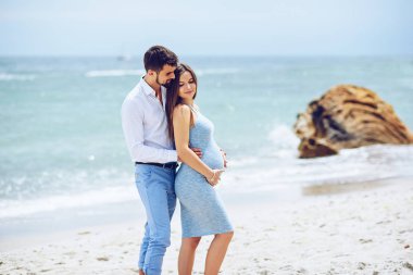 Beyaz bir gömlek ve Mavi pantolon cesur ve yakışıklı adam kayalar ve deniz arka planı mavi elbiseli güzel onun hamile eşi hugs.