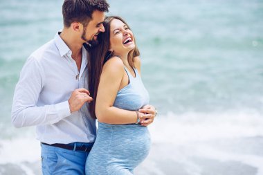Cesur ve yakışıklı adam beyaz bir gömlek ve Mavi pantolon deniz arka planı mavi elbiseli güzel onun gülümseyen hamile eşi hugs.