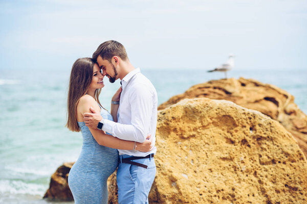 Красивая беременная женщина в синем платье и ее мужественный и красивый муж в рубашке и синих брюках прислонились друг к другу на фоне скалы и моря
.